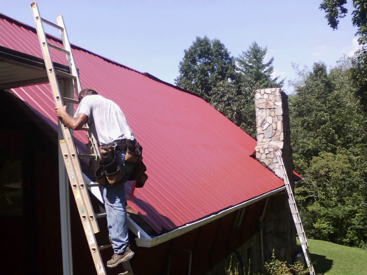 Roofer on a ladder beside a red metal roof and stone chimney — Soffit & Fascia Repair by Roofing Brothers in Steeles Tavern